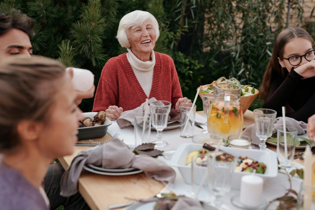 Una mujer mayor sonriente, con cabello canoso y un suéter rojo, disfruta de una comida al aire libre rodeada de otras personas más jóvenes, todos sentados alrededor de una mesa festiva con platos, vasos y jarras de bebida.