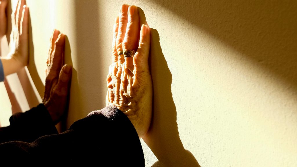 Primer plano de las manos de una persona mayor, apoyadas sobre una pared iluminada por la luz del sol, mostrando arrugas y detalles de envejecimiento. A su lado, se ve la mano de una persona más joven también apoyada sobre la pared.