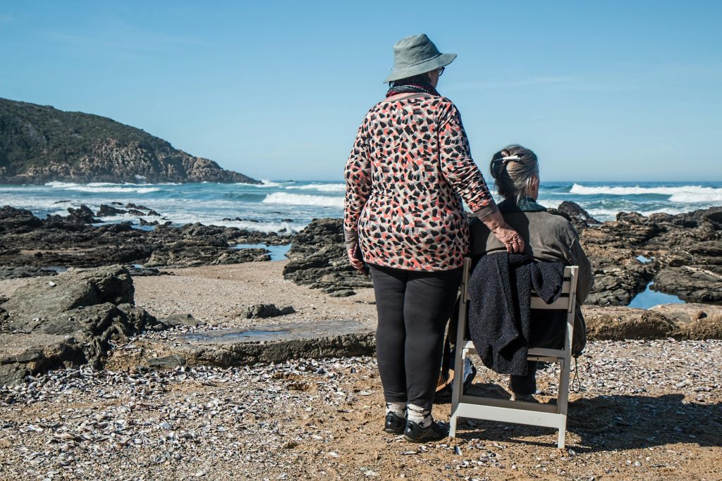 Dos mujeres mayores, una de pie y la otra sentada en una silla, disfrutan de la vista del mar en una playa rocosa. La mujer de pie tiene una mano en el hombro de la mujer sentada, mostrando un gesto de cuidado y compañía.